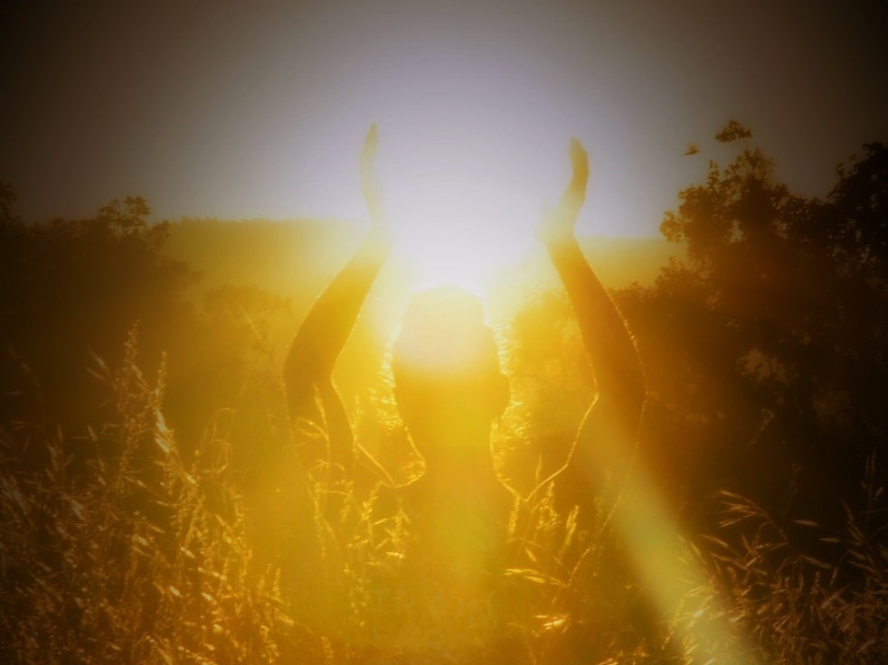 Image of a women in a field holding her arms up towards the sun. The light is very bright and golden.